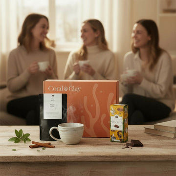 Lifestyle image featuring Coral & Clay For Friends gift box on a wooden table, styled with a steaming ceramic mug, cinnamon sticks, fresh mint leaves, and gourmet treats including artisan coffee and biscuits, in a cozy living room setting.