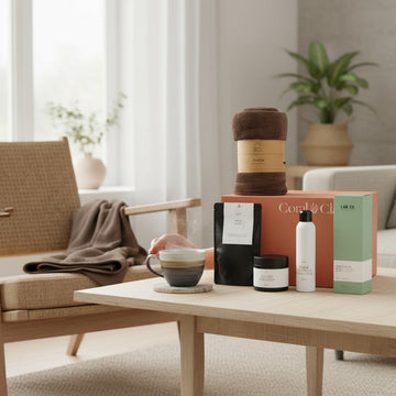 Coral & Clay grand gift box on a light wooden coffee table, styled with luxury self-care items including a folded brown towel, a black pouch, a white bottle, a black jar, and a green-packaged product, alongside a ceramic cup of coffee.