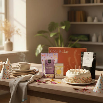 Birthday-themed lifestyle scene featuring Coral & Clay gift box with party hats, a birthday cake with candles, and premium treats including Best Birthday Cake mix and artisan coffee, styled on a wooden table in a cozy sunlit room.