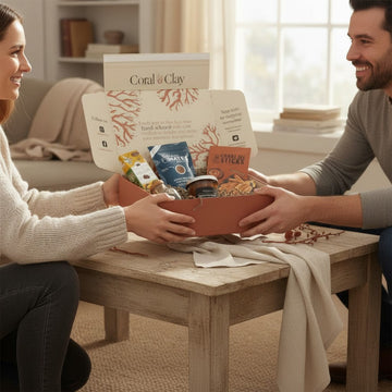 Lifestyle image featuring Coral & Clay thank-you gift box on a wooden coffee table, filled with premium treats including caramel sticks, artisan snacks, and gourmet items, in a cozy living room setting with natural light.
