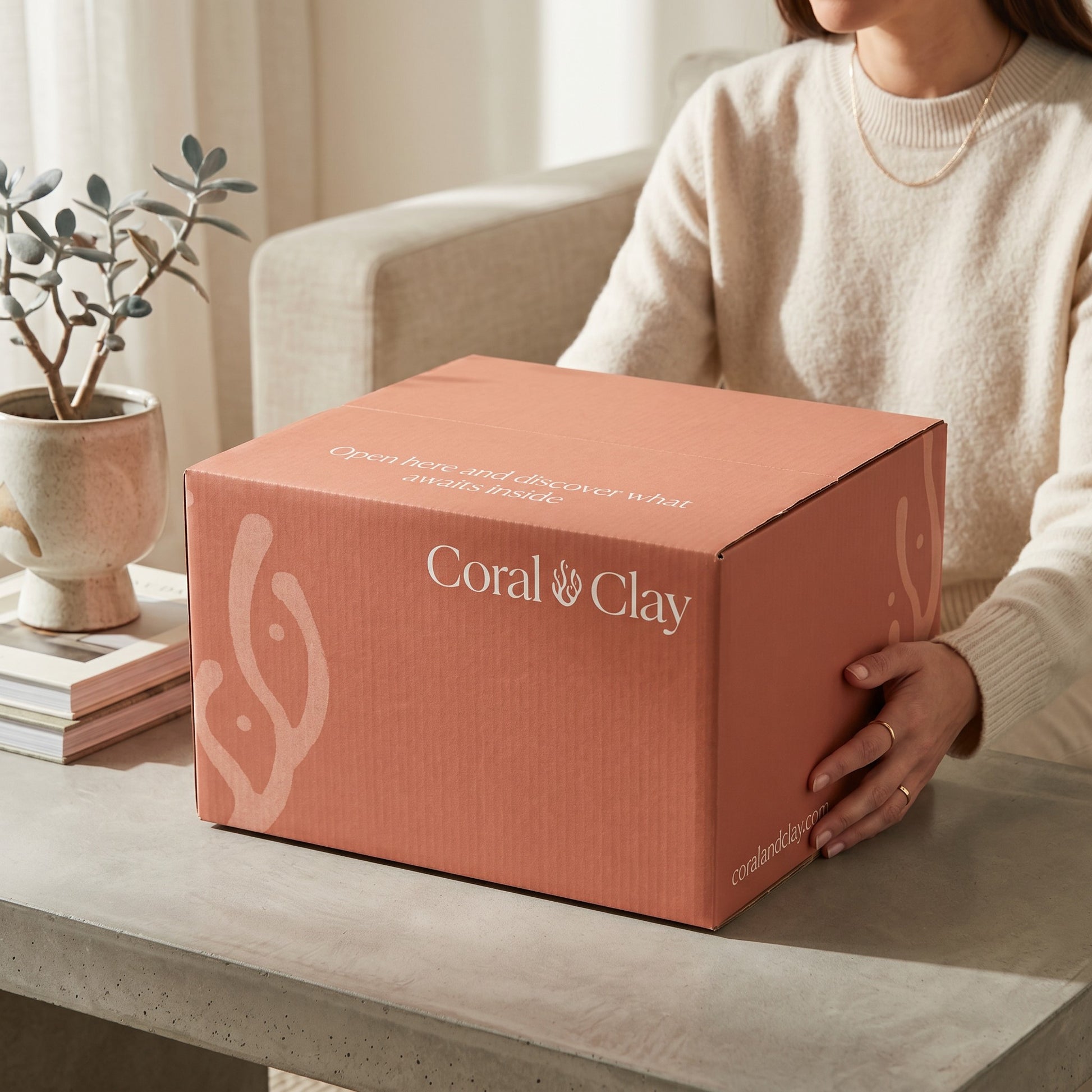 Person holding a Coral Clay Grand Gift box on a table with a plant and books in the background