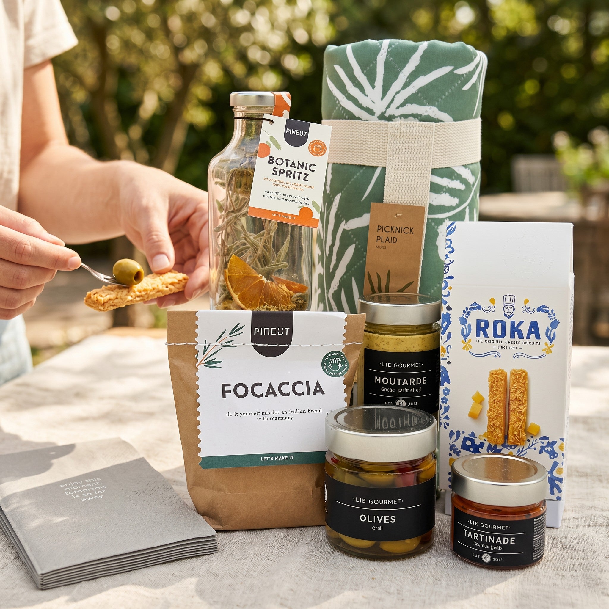 Gourmet Picnic Grand Gift picnic blanket setup with focaccia bread mix, grilled red pepper tapenade, basil parsley garlic mustard, chili olives, Gouda cheese sticks, botanic spritz mocktail, napkins and picnic blanket.