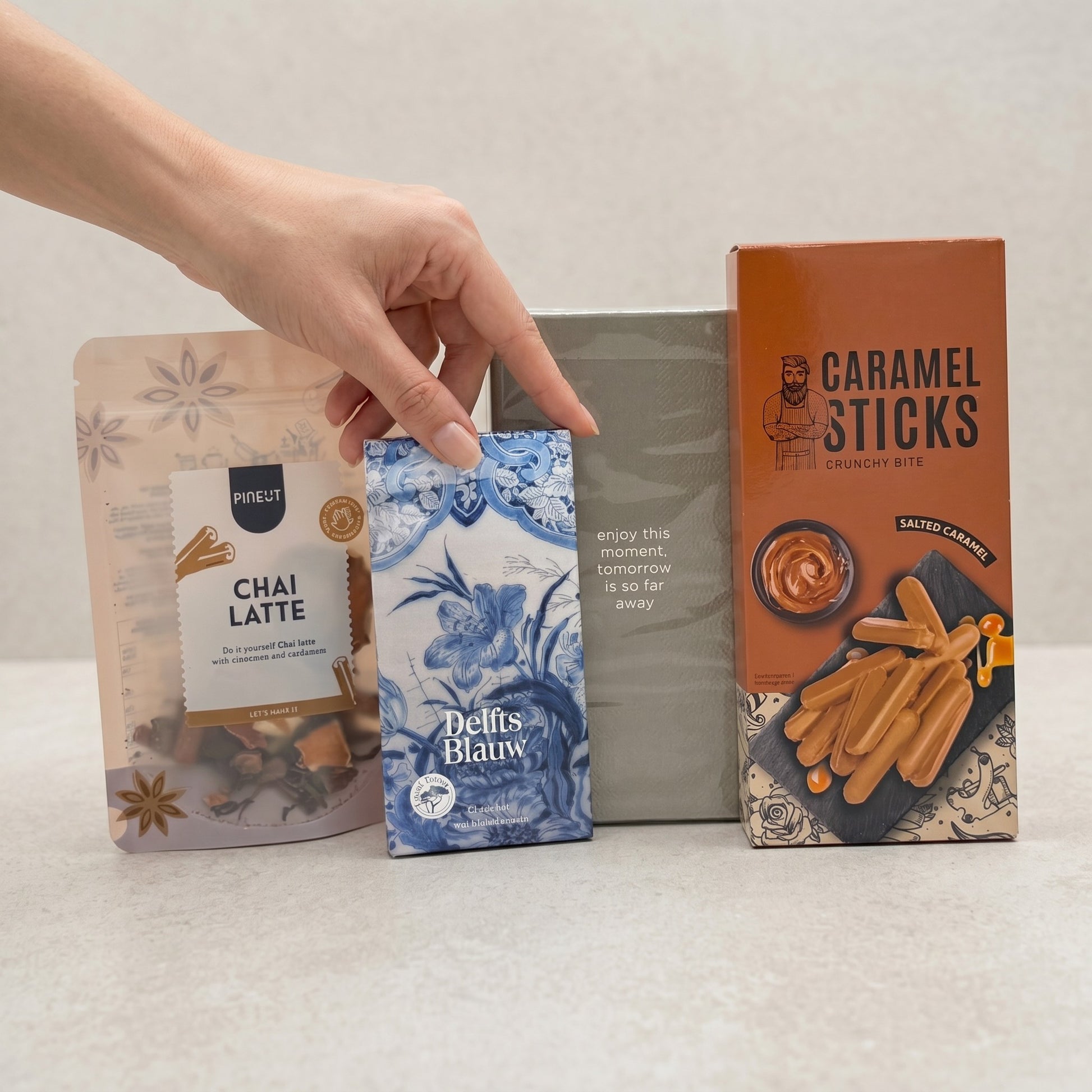 Coral & Clay Sunday Afternoon Chai & Chocolate letterbox gift photo – hand reaching for Delfts Blauw blueberry chocolate bar with chai and salted caramel treats on a white surface.
