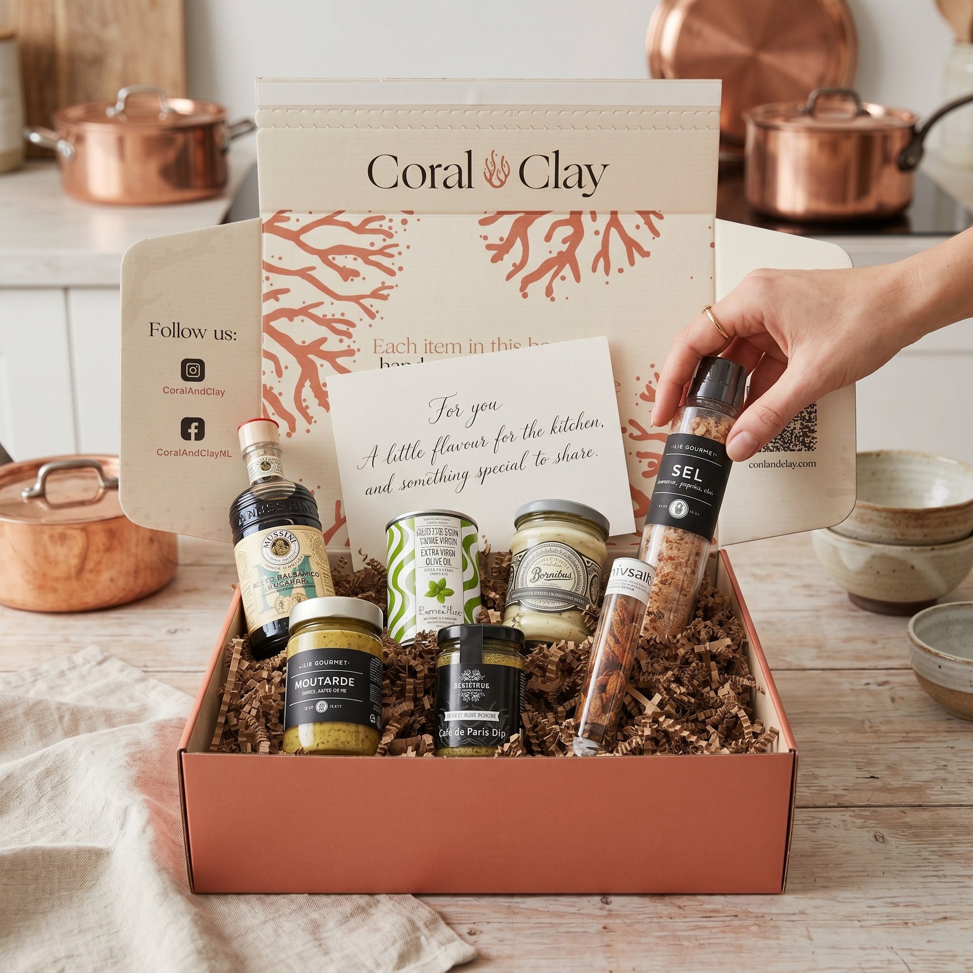 Gift box with various items including bottles and jars, with a hand reaching in to take one, set against a kitchen background.