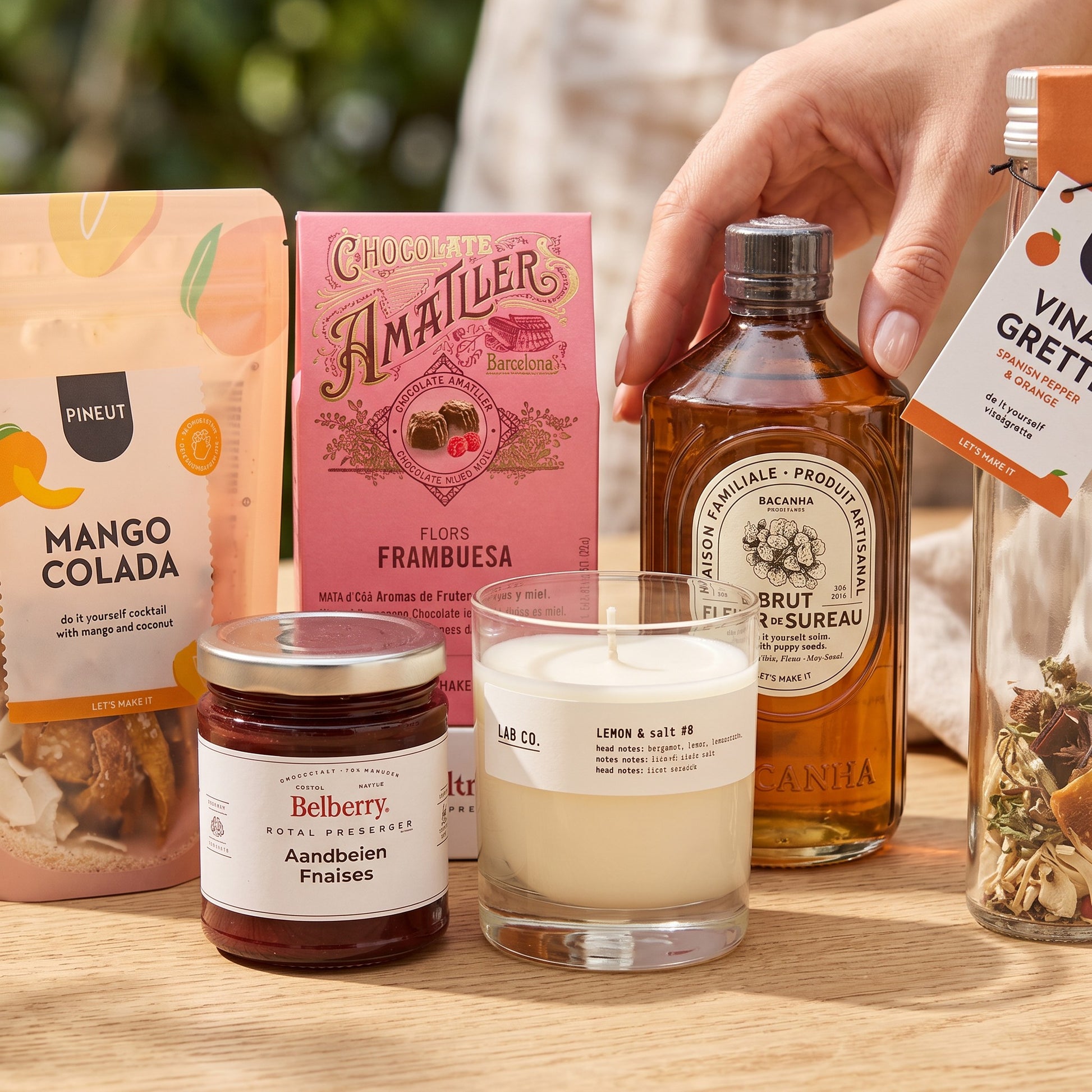 Coral & Clay Summer Feeling Signature Gift close-up photo – table close-up showing Mango Colada drink mix, elderflower syrup, lemon & poppy cake mix, strawberry confiture, raspberry pralines, Spanish pepper & orange vinaigrette, and scented candle.
