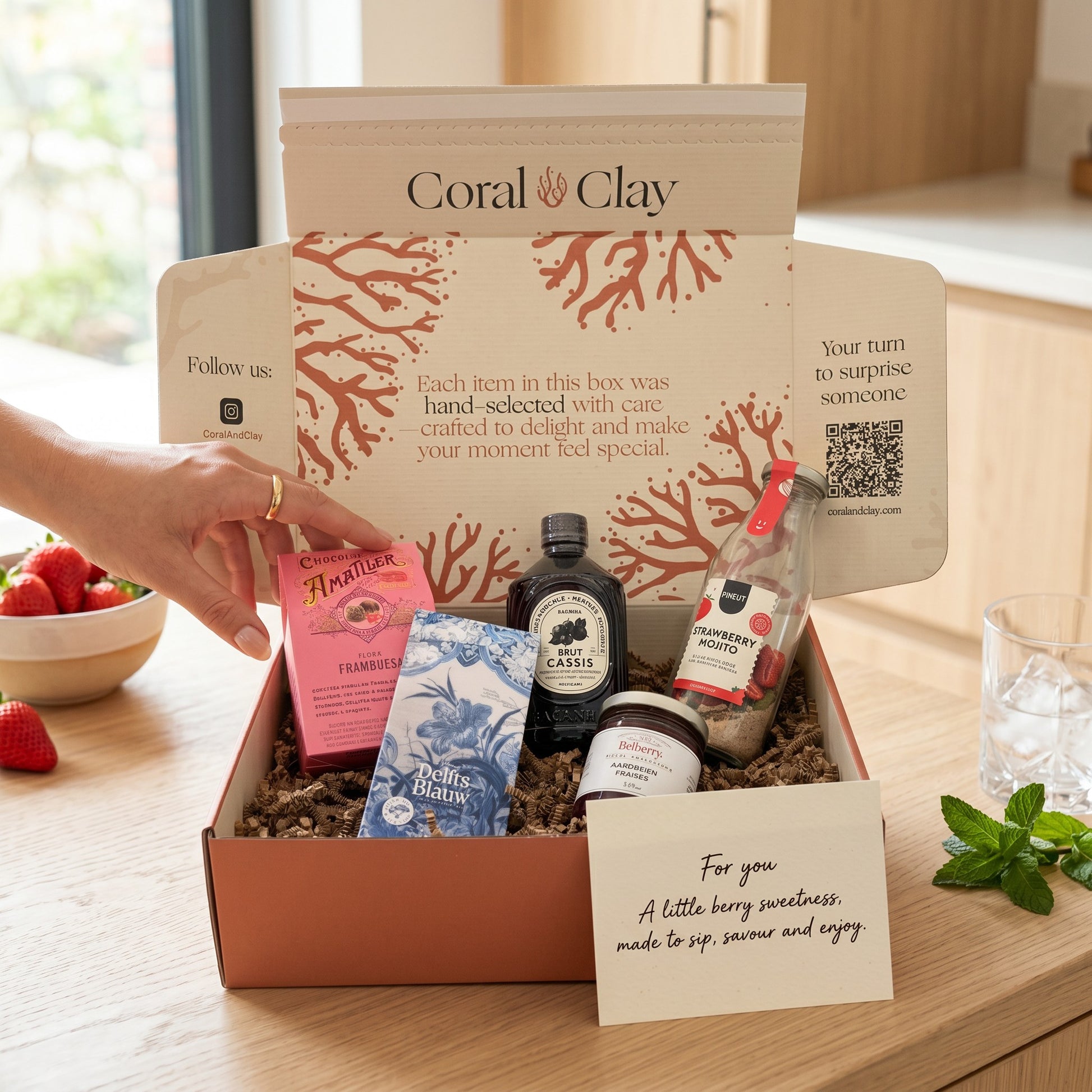 Coral & Clay Very Berry Signature Gift staged kitchen shot with open box showing Amatller milk chocolate raspberry pralines, Delfts Blauw blueberry chocolate bar, Bacanha brut cassis syrup, Belberry strawberry jam, Pineut 0% Strawberry Mojito bottle and handwritten gift note card.