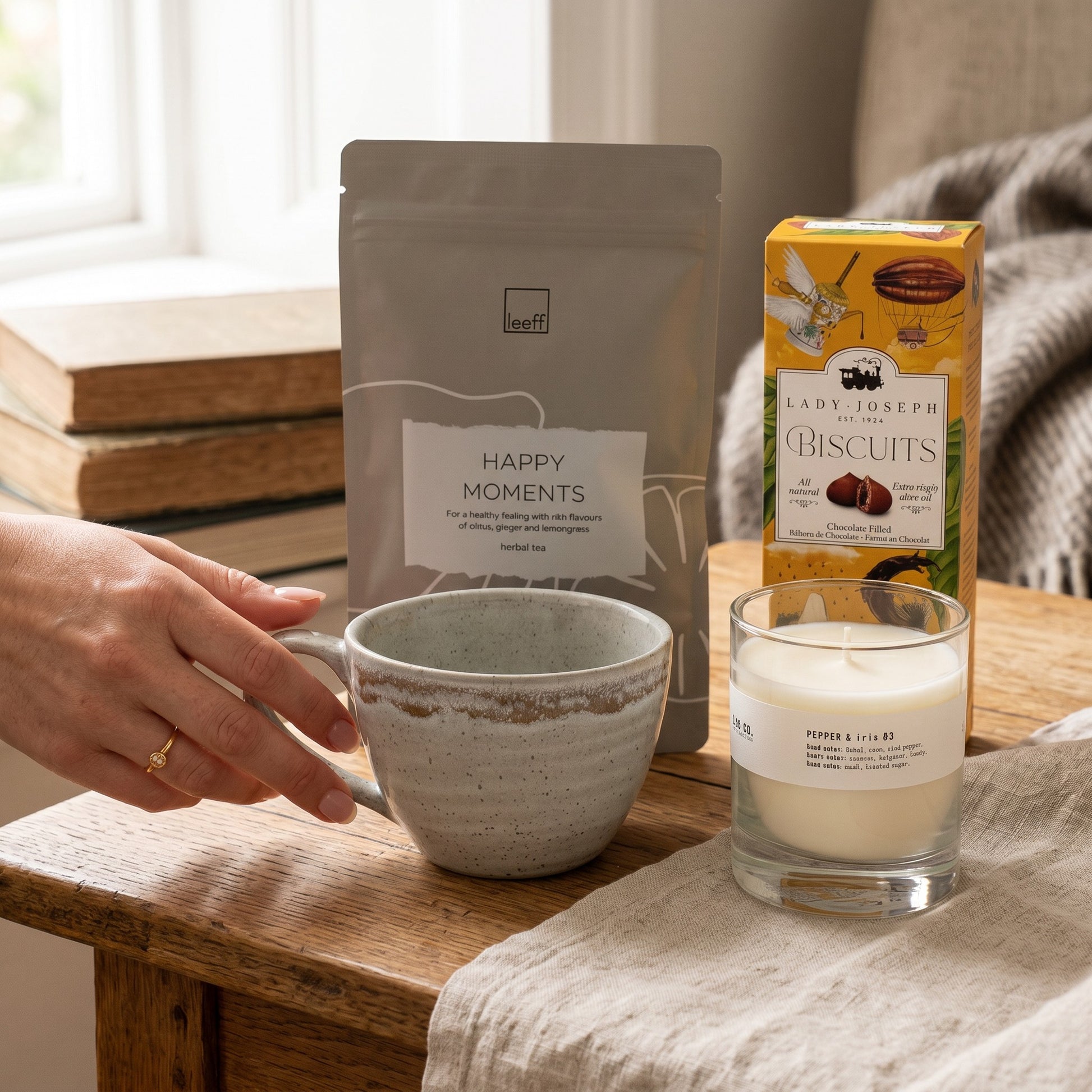Person holding a mug with coffee, a bag of 'Happy Moments' coffee, a box of 'Lady Joseph Biscuits', and a candle on a wooden table.