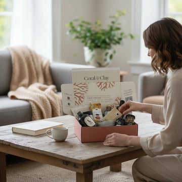 Woman opening a Coral & Clay gift box on a wooden table in a cozy living room.