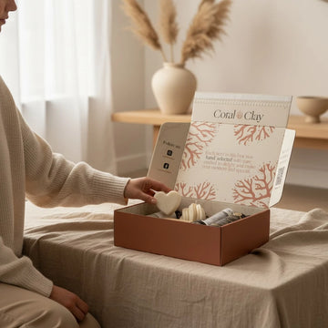 Person opening Coral & Clay signature gift box with decorative items inside, in a cozy indoor setting.