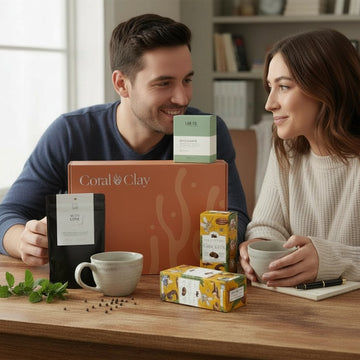 Coral & Clay Anniversary gift box displayed on a wooden table with artisanal treats and tea essentials, including loose-leaf tea, premium biscuits, and elegant ceramic mugs. Fresh mint leaves and a notebook add a cozy, intimate touch to the setting.
